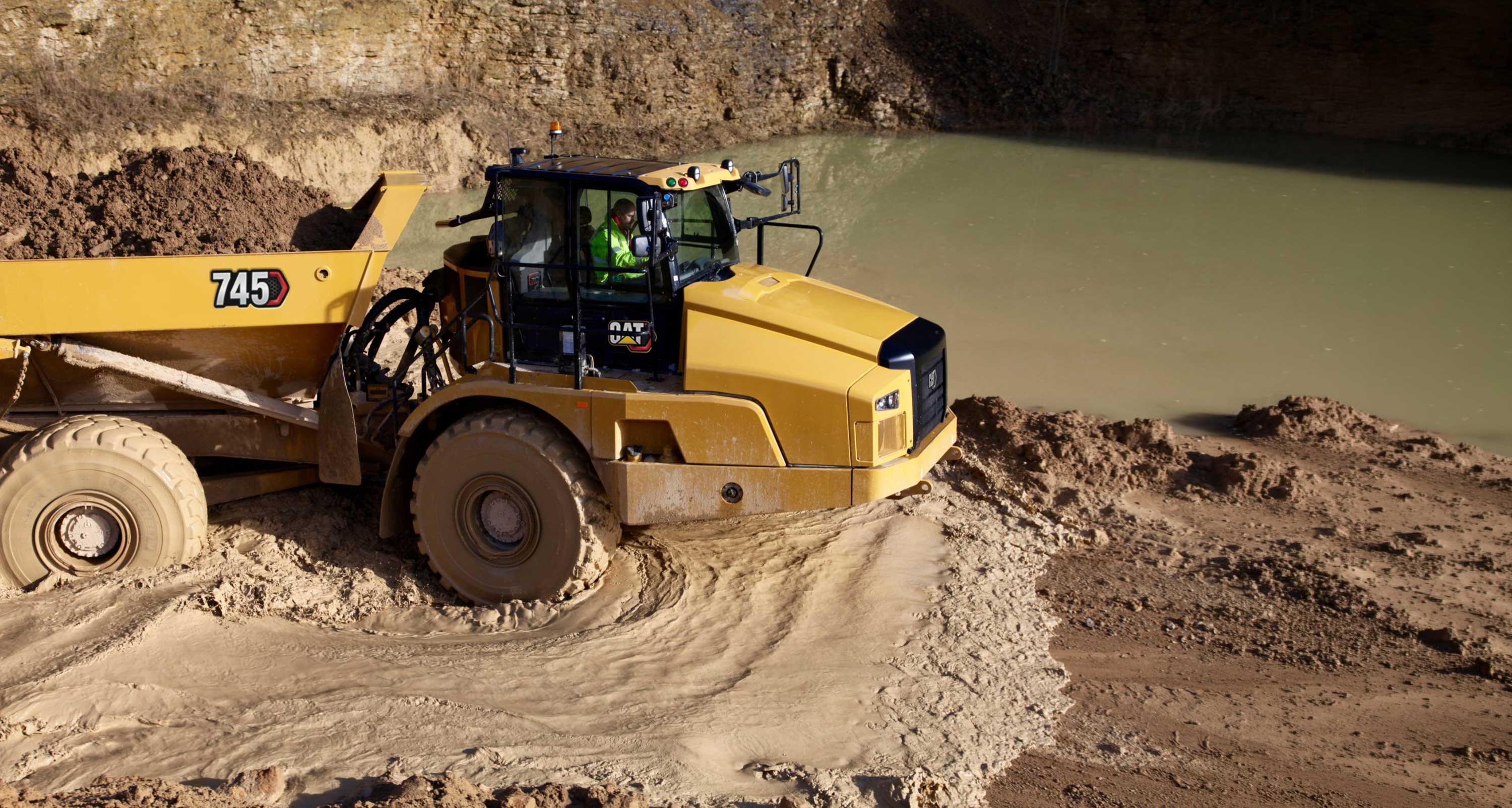 Cat 745 articulated dump truck driving through a muddy excavation site beside a large water-filled pit.